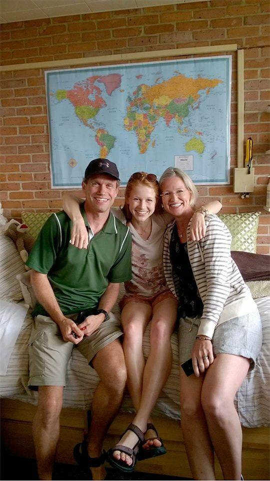Samantha Hilburn with her arms around her parents sitting on the bed in her dorm room