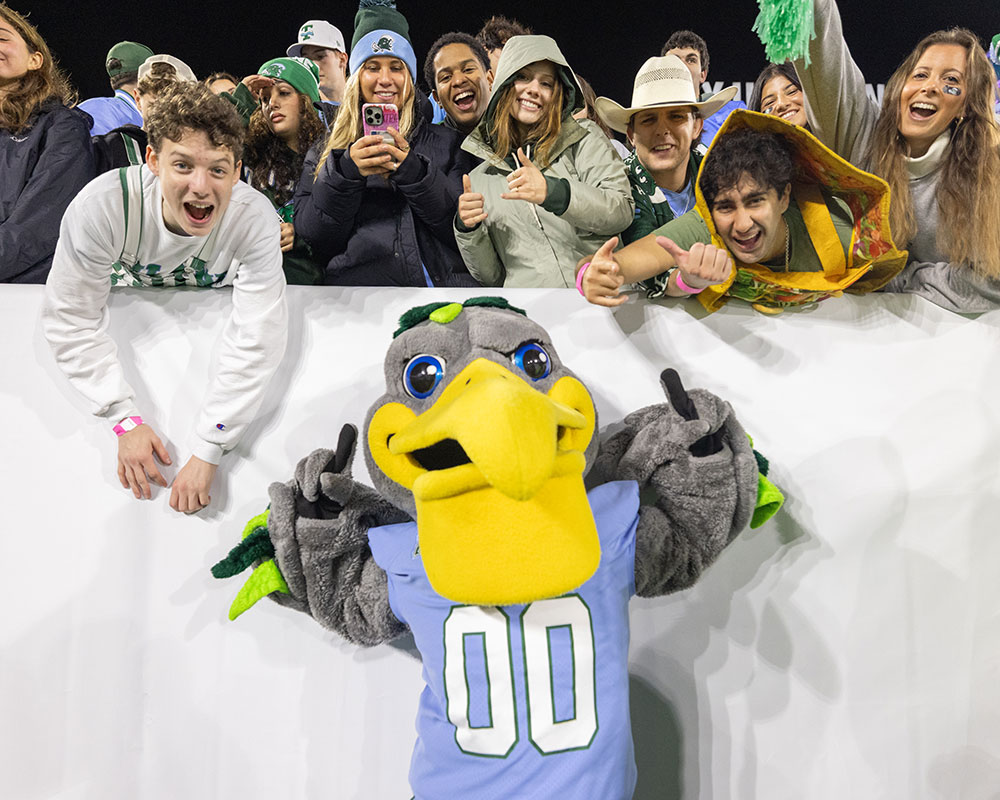 Riptide the mascot gives thumbs-up, with cheering fans at a football game