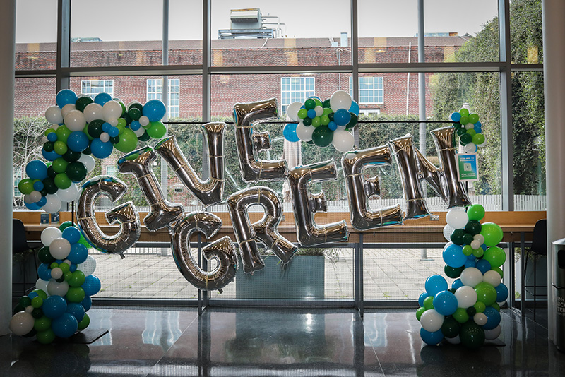 Silver "GIVE GREEN" balloons framed by green, blue, and white balloon arches.