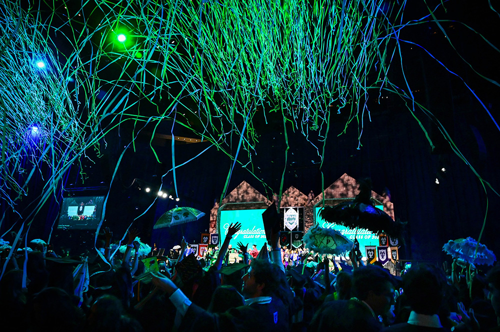 Graduates celebrating under falling green and blue confetti at 2026 commencement