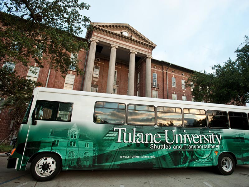 A Tulane shuttle bus is pictured parked in front of Newcomb Hall with a bright green Tulane University shuttle bus wrap covering its sides.
